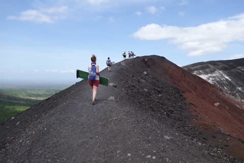 A hiker carrying a green board along the black ridge, with the red crater slope to the right and others ahead