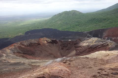 Looking into the reddish-brown crater with steam rising, black lava fields and green hills beyond