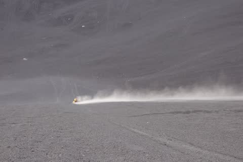 A boarder speeding down the grey ash slope, trailing a long plume of dust