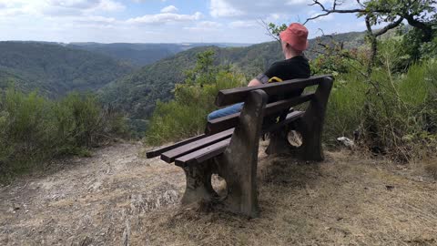Someone in a sun hat sitting on a bench at the edge of the slope, looking out over the wooded Lahn Valley