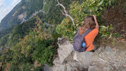A hiker with a backpack among rocks and bushes on a steep slope, with a village in the valley far below