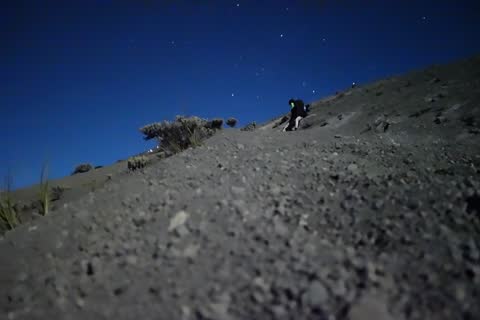 A hiker with a headlamp climbing a steep slope of volcanic scree at night under the stars