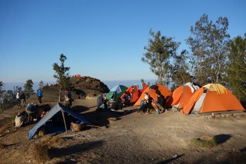 Orange tents and hikers at the camp on the crater rim in the evening sun, above a sea of clouds